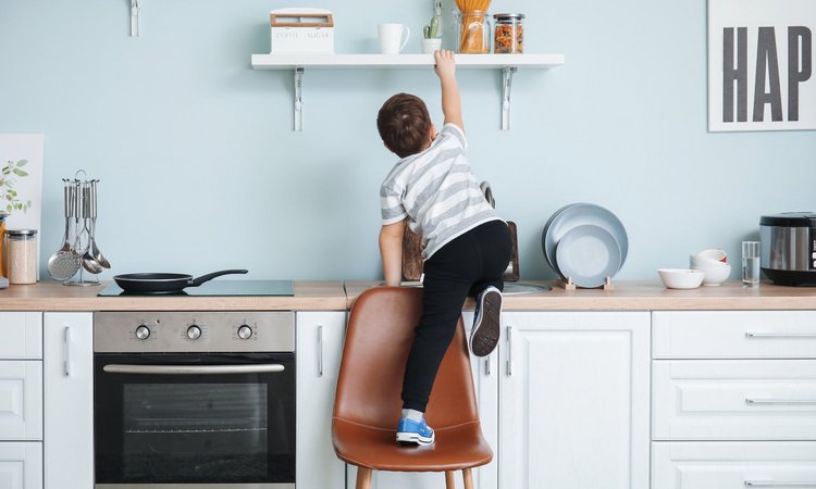 Ein Kind klettert über einen Stuhl auf die Küchenzeile um an ein Regal zu kommen