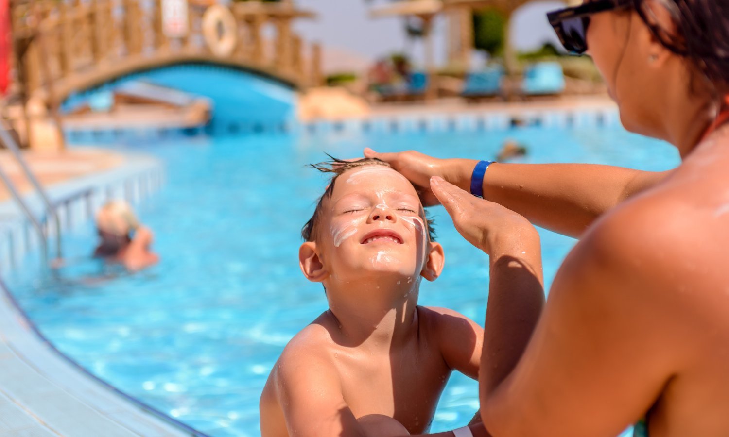Eine Frau cremt ein Kind bei strahlendem Sonnenschein am Pool im Gesicht mit Sonnencreme ein