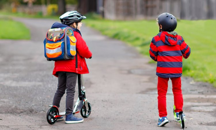 Zwei Kinder mit Helm fahren mit dem Roller über einen Feldweg