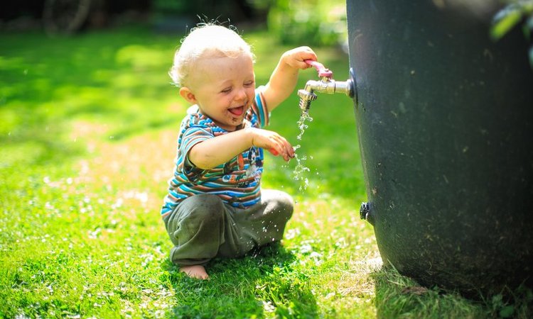 Ein Kleinkind spielt fröhlich mit dem Wasser aus der Regentonne