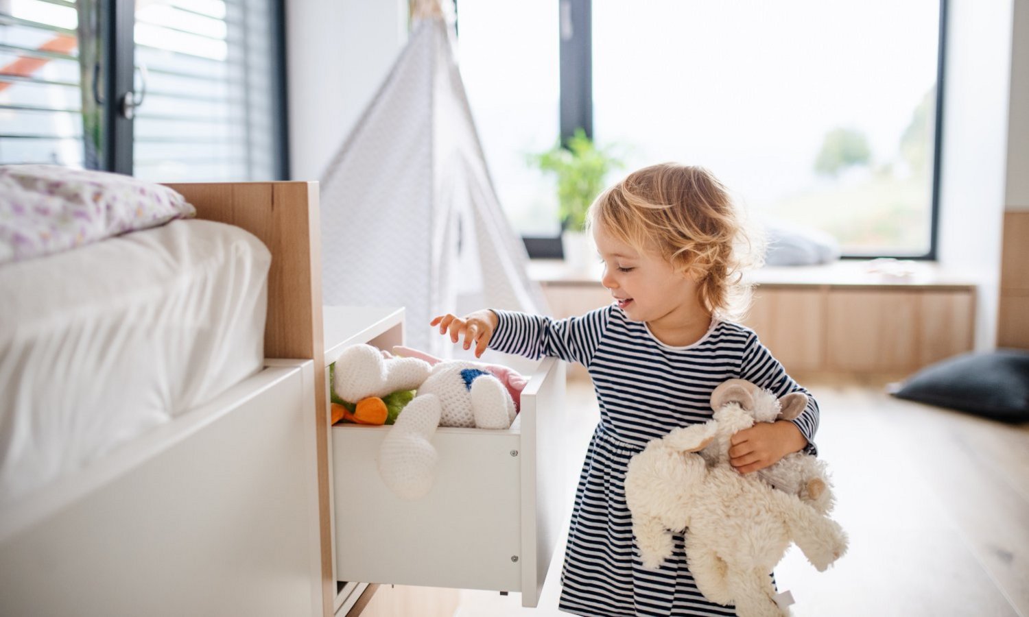 Ein Kleinkind räumt Stofftiere aus einer Schublade im lichtdurchfluteten Kinderzimmer