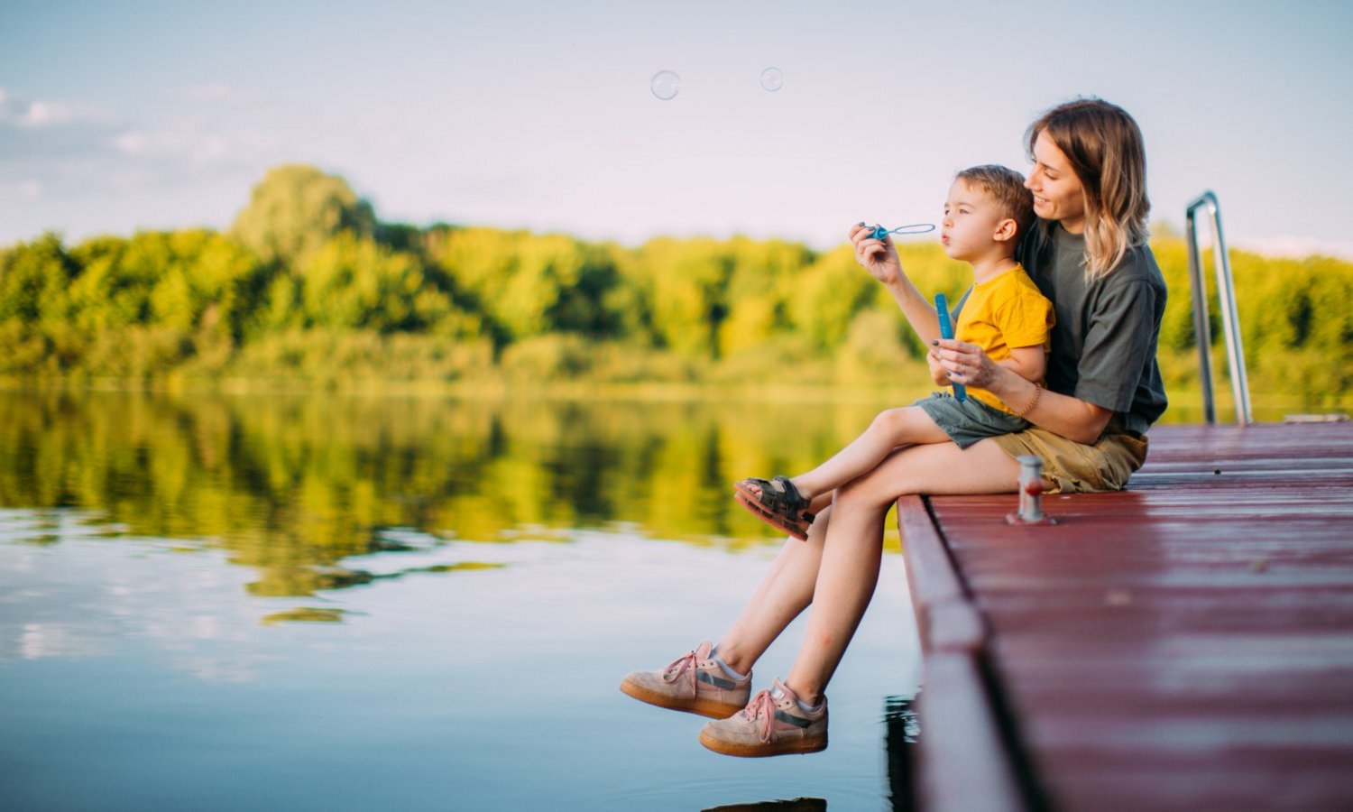 Eine Frau und ein Kind sitzen am Steg eines Sees und blasen Seifenblasen