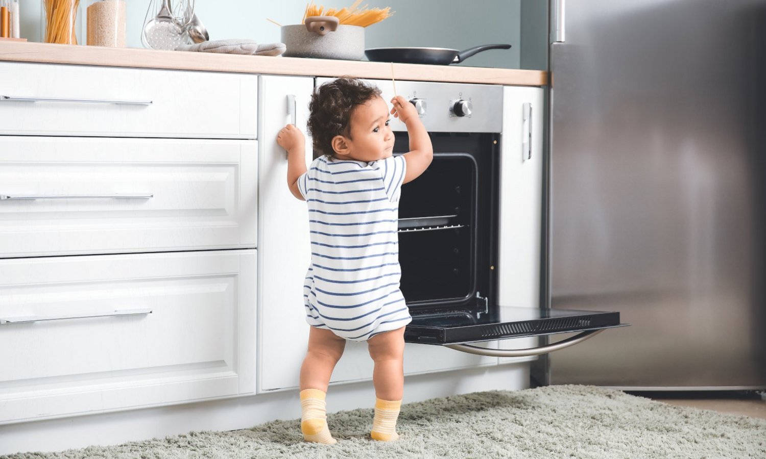 Ein Kleinkind steht vor der weißen Küchenzeile am geöffneten Backofen und greift nach den Nudeln auf dem Herd