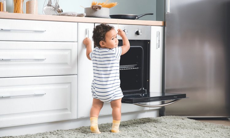 Ein Kleinkind steht vor der weißen Küchenzeile am geöffneten Backofen und greift nach den Nudeln auf dem Herd