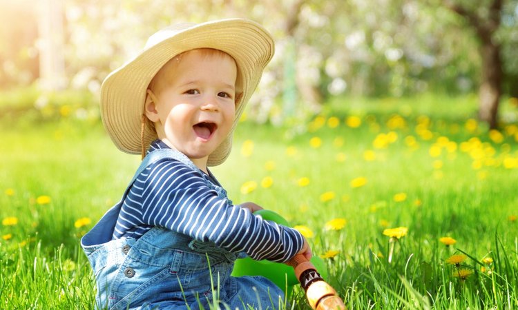 Ein Kleinkind mit Sonnenhut sitzt bei Sonnenschein auf einer Wiese und spielt mit dem Gartenschlauch