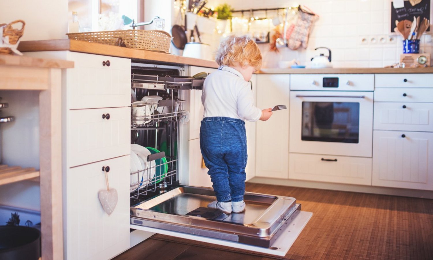 Ein Kleinkind steht auf der aufgeklappten Spülmaschinentürn in der Küche