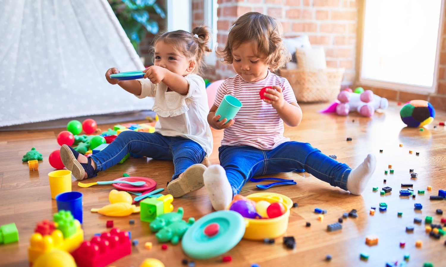 Zwei Kleinkinder sitzen inmitten von buntem Spielzeug und Kleinteilen