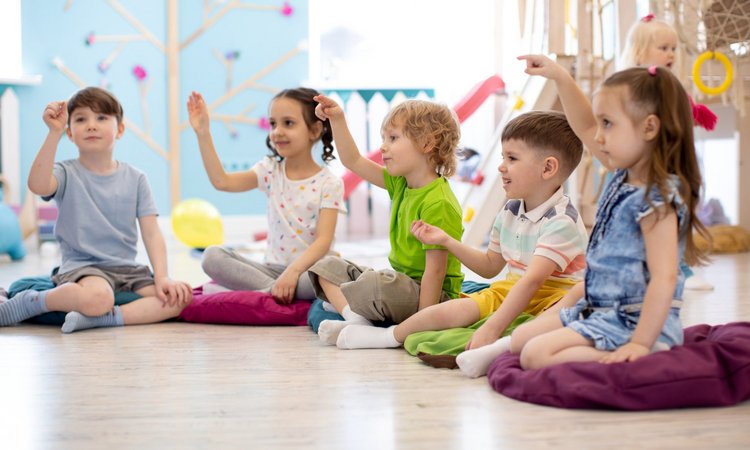 Kinder sitzen im Halbkreis auf dem Fußboden und zeigen auf