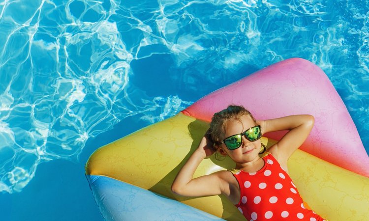 Ein Kind mit Sonnenbrille und Badeanzug liegt bei Sonnenschein entspannt auf einer Luftmatratze im Pool